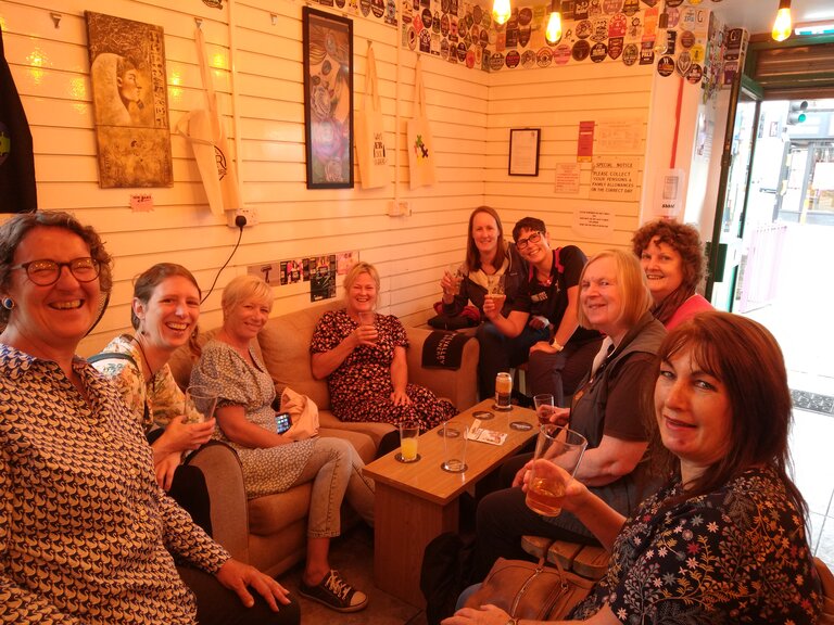 A group of women sitting round a low table smile and raise their beer glasses to the camera.